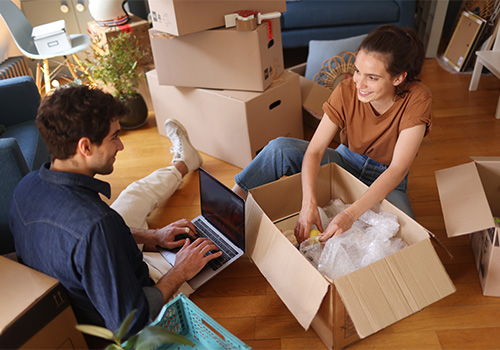 A couple organising moving boxes at home and planning cargo van rental on a laptop after moving to Portugal.