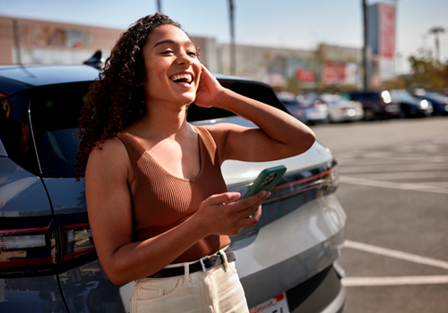 Smiling young woman, leaning on her car while holding her phone