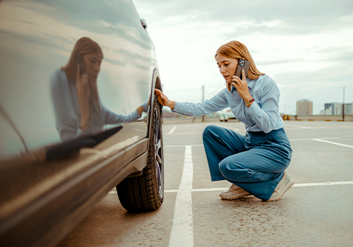 Driver crouching next to the tire of a broken-down car, talking on the phone to request roadside assistance in a parking lot.