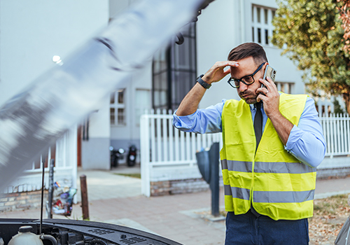 Driver wearing a reflective vest calling roadside assistance next to a broken-down car with the hood open on a residential street.