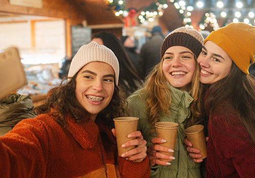  Três raparigas divertem-se num mercado de inverno, enquanto tiram uma selfie e bebem chocolate quente. 