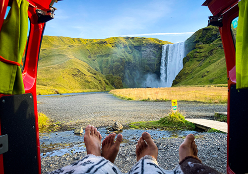  Traseira de carrinha aberta e ocupantes sentados de frente para uma cascata na Islândia. 