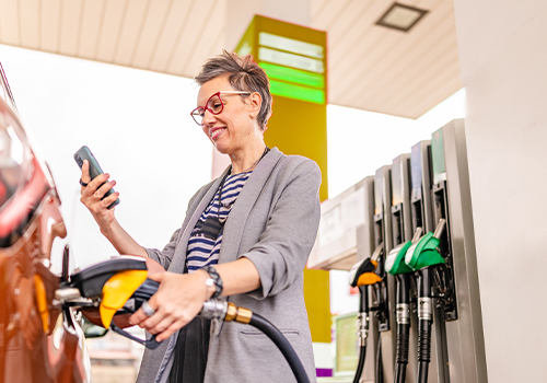 Driver refueling her car at a gas station while using an app to estimate fuel cost for a trip.