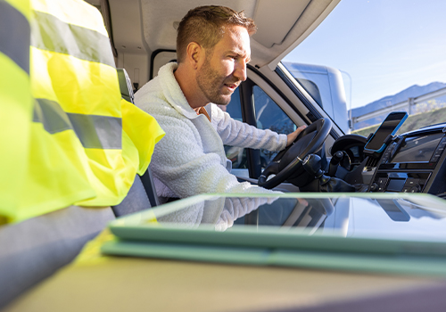 Man in the driver’s seat of a van, with a smartphone mounted on the dashboard, leaning forward to adjust something inside the vehicle; a high-visibility vest is on the seat beside him.