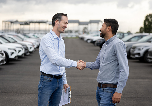Dois homens a apertar as mãos num parque de estacionamento cheio de carros, sugerindo acordo ou entrega de veículo.