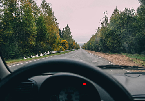 Vista do interior de um carro com uma luz vermelha acesa e com o volante em primeiro plano, com estrada aberta à frente.