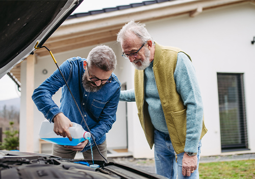 Driver checking the engine oil level with the hood open while another person observes during a car maintenance check at home.