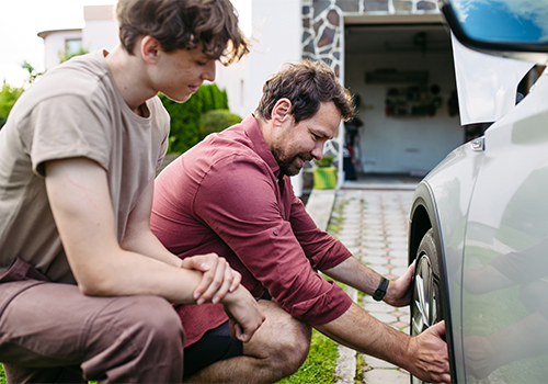Driver checking the engine oil level with the hood open while another person observes during a car maintenance check at home.