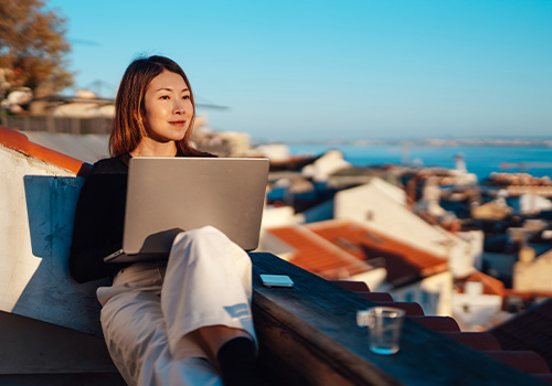 Young Asian woman working on a laptop, sitting on a rooftop under a clear blue sky