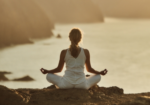 Woman seated in lotus position on a cliff above the sea, seen from behind, meditating in silence.