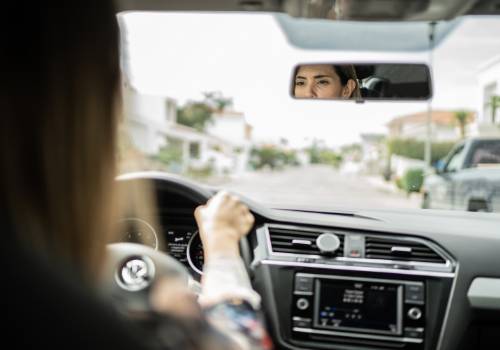 of a woman driving, seen in the car’s rear-view mirror